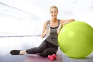 smiling woman sitting next to fitness ball and holding in hand a bottle of water while relaxing after fitness workout